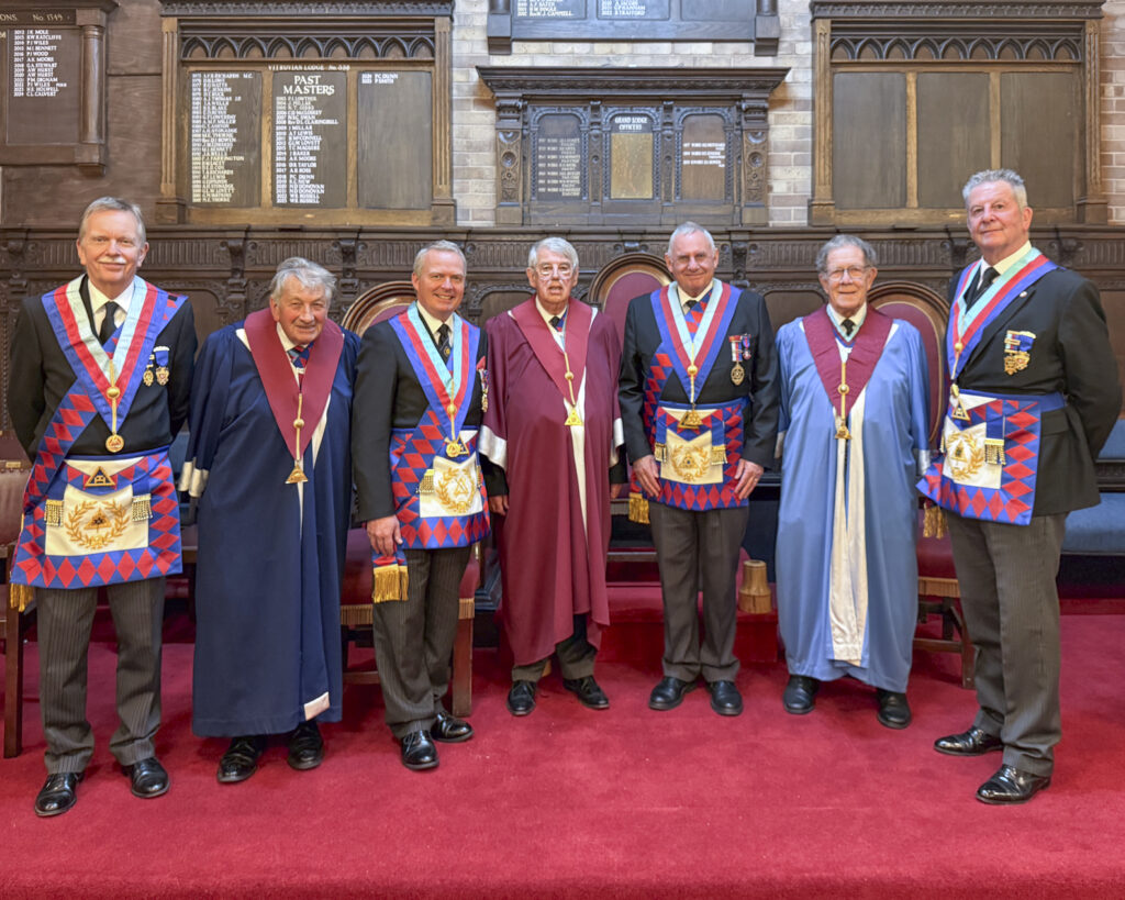 A group of seven royal arch masons standing side-by-side in a formal setting, wearing dark suits and royal arch regalia. They are positioned in front of a wooden wall with framed lists of names.