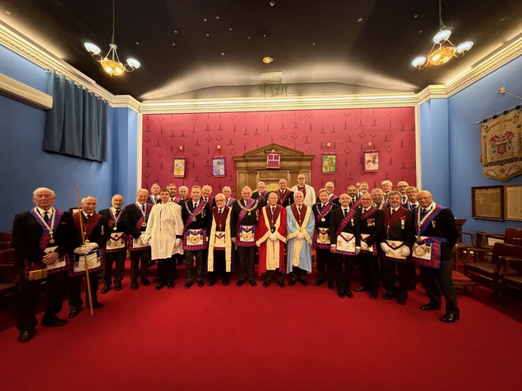 A group of men in formal Royal Arch regalia, including aprons, collars, and chains, standing together in a room with red walls and blue curtains.