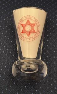 A Firing glass created for the 1000th meeting of Palladian chapter featuring a gold, six-pointed star emblem with a central triangle, surrounded by the Latin text "DEO - REGI - FRATRIBUS - HONOR - FIDELITAS - BENEVOLENTIA" and the English words "WISDOM, PEACE, STRENGTH, CONCORD, BEAUTY, TRUTH."
