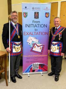 Two men in Masonic regalia, including blue and red aprons and collars, standing on either side of a pull-up banner that reads "From INITIATION to EXALTATION" with images of Masonic aprons and a QR code.