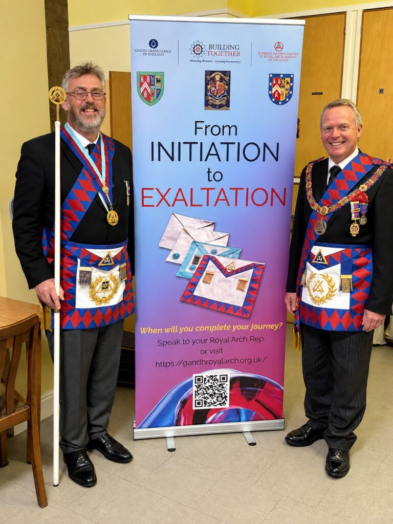 Two men in Masonic regalia, including blue and red aprons and collars, standing on either side of a pull-up banner that reads "From INITIATION to EXALTATION" with images of Masonic aprons and a QR code.
